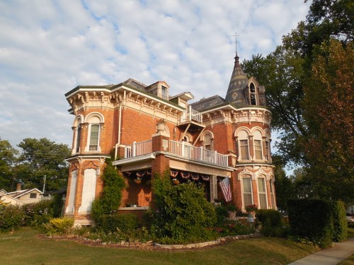 Victorian-style brick house with a turret, balcony, and lush greenery, set against a cloudy sky.