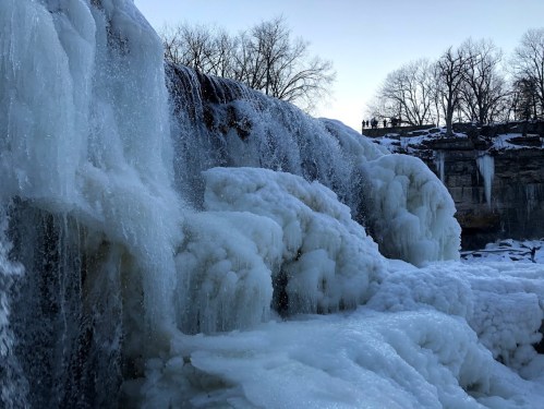 A frozen waterfall with icy formations, surrounded by trees and a clear sky in the background.