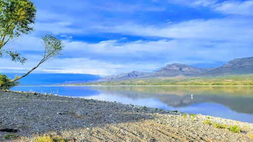 A serene lakeside view with mountains in the background, under a blue sky with scattered clouds.