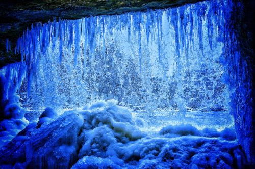 A frozen waterfall with icicles and blue-tinted water cascading over rocks, creating a serene winter scene.