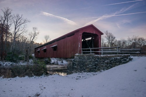 A red covered bridge stands over a snowy landscape, surrounded by bare trees and a calm river under a clear sky.