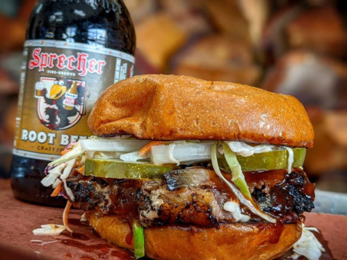 A close-up of a juicy burger with pickles and onions, alongside a bottle of Sprecher root beer.