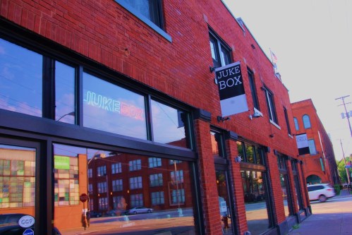 Exterior of a brick building featuring a sign that reads "JUKE BOX" and large windows reflecting the street.