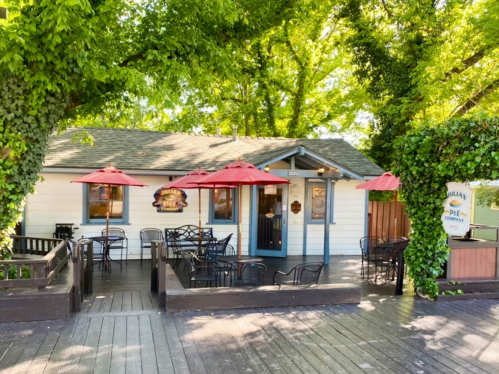 A charming white cottage with a porch, red umbrellas, and outdoor seating surrounded by greenery.