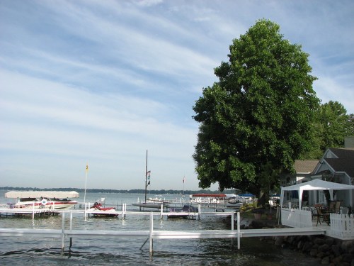 A serene lakeside scene with boats docked, a large tree, and a white fence under a blue sky with wispy clouds.