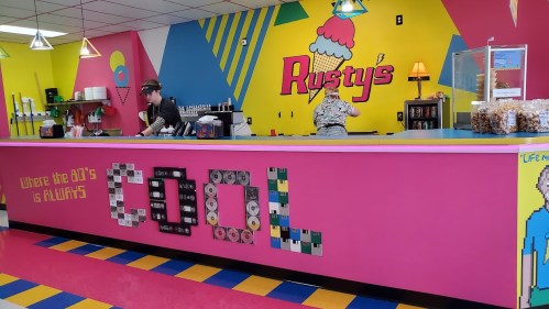 A colorful ice cream shop interior with a vibrant pink and yellow wall, featuring retro decor and a counter with staff.