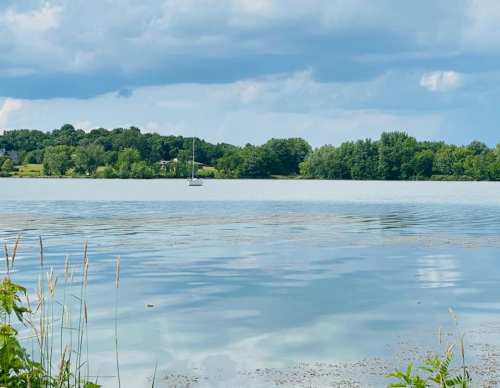 A calm lake with a sailboat in the distance, surrounded by lush greenery and a cloudy sky.