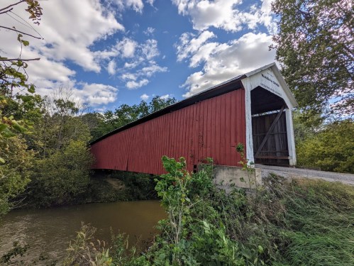 A red covered bridge spans a calm river, surrounded by greenery and under a partly cloudy sky.