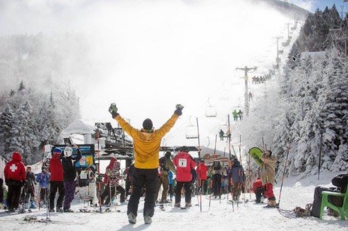 A crowd of skiers and snowboarders at a snowy mountain resort, with a person in a yellow jacket celebrating.