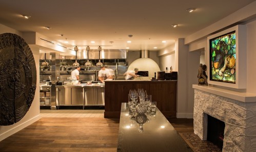 A modern restaurant interior featuring a kitchen, stone fireplace, and stained glass artwork.