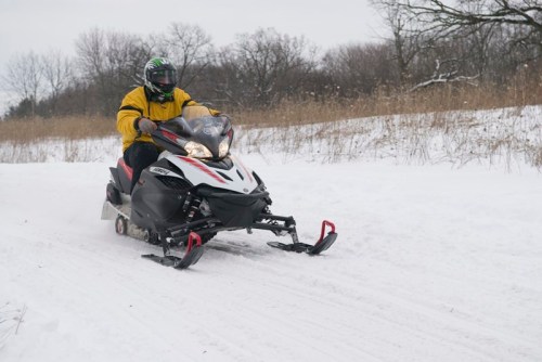 A person in a yellow jacket rides a snowmobile on a snowy trail surrounded by bare trees and grass.