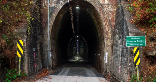 A dark, arched tunnel with brick walls, featuring a sign for the historic Dingess Tunnel and a paved road inside.