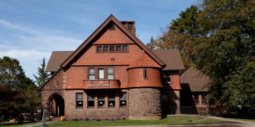 A large, historic brownstone building with a steep roof and decorative brickwork, surrounded by trees and flowers.