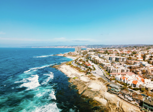 Aerial view of a coastal city with waves crashing on rocks and buildings lining the shore under a clear blue sky.