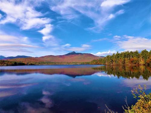 A serene lake reflects a vibrant autumn landscape with mountains and a blue sky dotted with clouds.