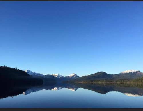 A serene lake reflects snow-capped mountains under a clear blue sky, surrounded by lush greenery.