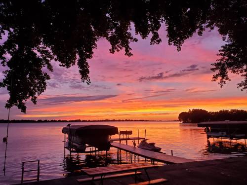 A serene lake at sunset, with vibrant pink and orange skies reflecting on the water and silhouettes of boats and trees.