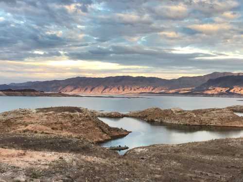 A serene landscape featuring a calm lake surrounded by rocky hills and mountains under a cloudy sky.