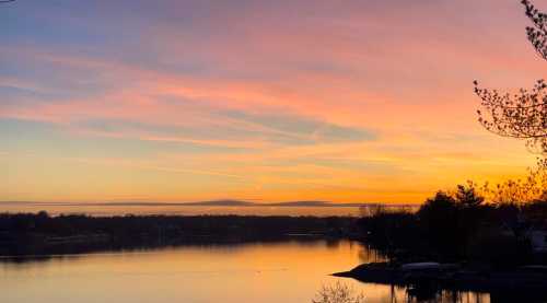 A serene sunset over a calm lake, with vibrant colors reflecting on the water and silhouettes of trees in the foreground.