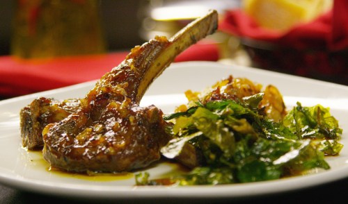 A plate featuring a lamb chop with a savory glaze, accompanied by sautéed greens and crispy potatoes.