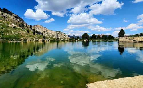 A serene lake reflecting clouds and mountains, surrounded by greenery under a bright blue sky.