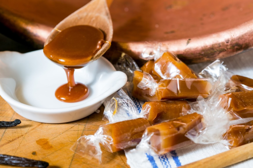 A wooden spoon drizzles caramel sauce over a white dish, with wrapped caramel candies scattered on a wooden surface.