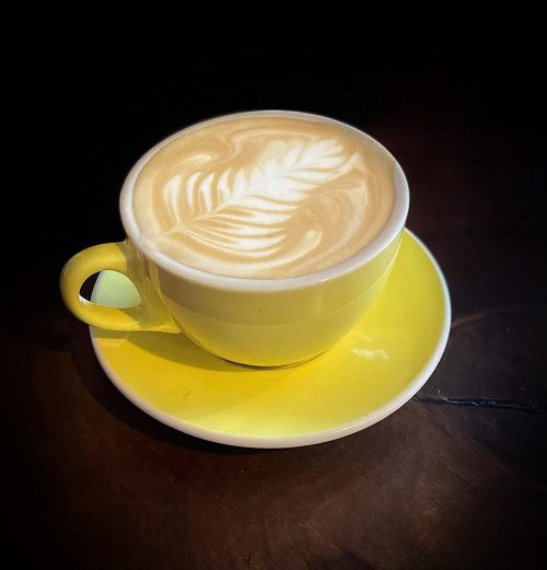 A yellow cup of latte art featuring a leaf design, sitting on a matching saucer against a dark background.