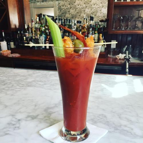A tall glass of Bloody Mary garnished with celery, shrimp, and olives, set on a marble bar with a blurred background.