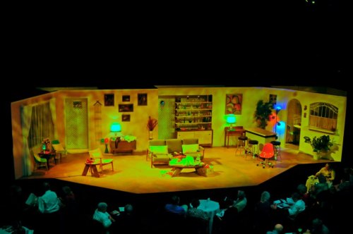 A brightly lit theater stage set with colorful furniture, plants, and a piano, viewed from the audience.