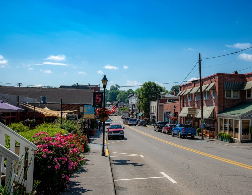 A charming small town street lined with shops, flowers, and parked cars under a clear blue sky.