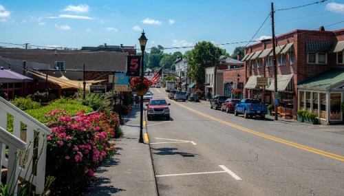 A charming small town street lined with shops, flowers, and parked cars under a clear blue sky.