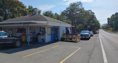 A small roadside eatery with a blue exterior, parked cars, and trees lining the street on a sunny day.