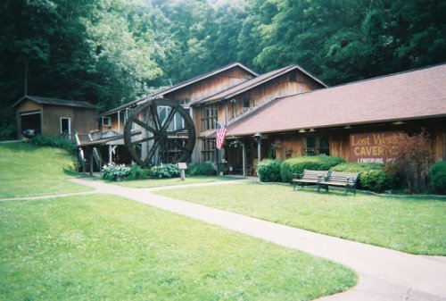 A rustic building with a water wheel, surrounded by green grass and trees, featuring an American flag and benches.