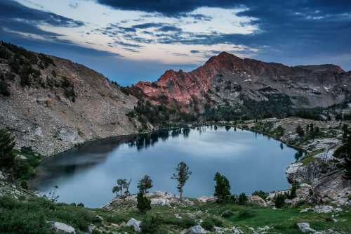 A serene mountain lake surrounded by rocky terrain and trees, reflecting the sky at dusk.