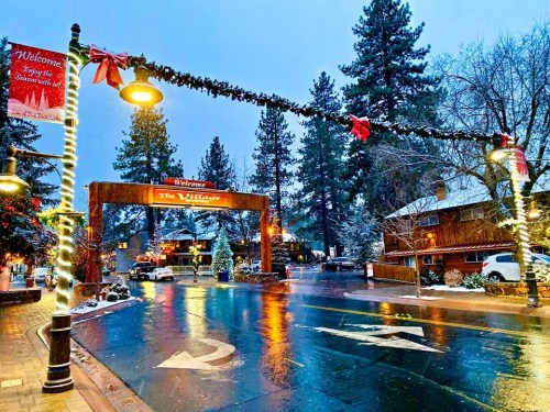 A snowy village entrance adorned with festive decorations and streetlights, surrounded by tall pine trees.