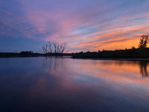 A serene river at sunset, reflecting vibrant pink and orange hues with silhouetted trees in the background.