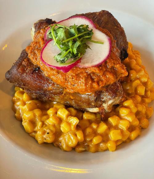 A plate featuring crispy chicken topped with a red sauce, garnished with radish and cilantro, served over creamy corn.