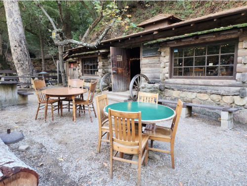 A rustic outdoor seating area with wooden tables and chairs near a log cabin in a forested setting.