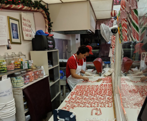 A candy shop worker shapes peppermint candies on a countertop, surrounded by jars and festive decorations.