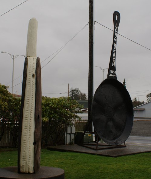 Two oversized sculptures: a giant wooden spoon and a large fork, set against a cloudy sky in Long Beach, Washington.