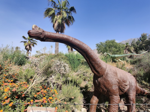 A large dinosaur statue with a long neck stands among colorful flowers and palm trees under a clear blue sky.