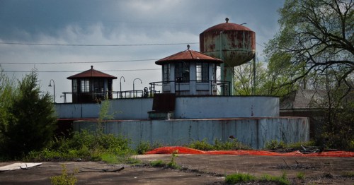 Abandoned industrial site featuring two water towers and overgrown vegetation under a cloudy sky.