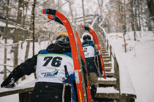Two athletes in winter gear climb snowy stairs, carrying sleds for a luge competition.