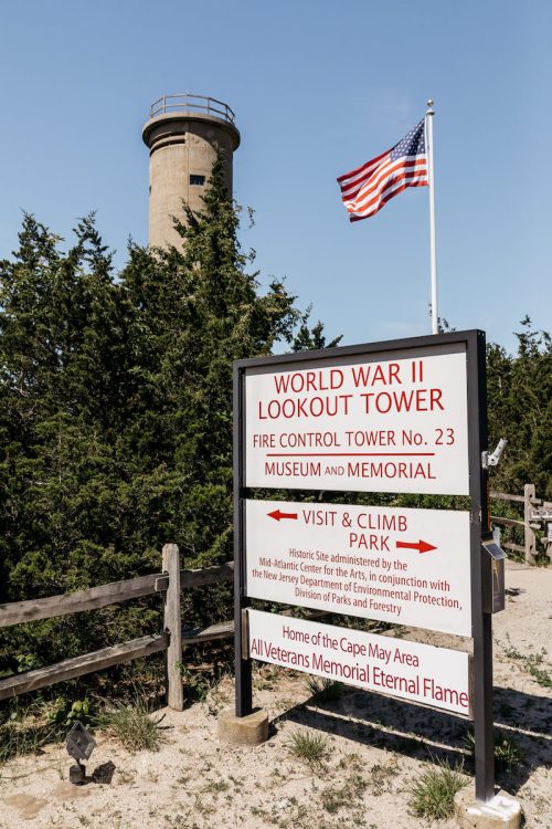 Sign for World War II Lookout Tower No. 23, with an American flag and a tower in the background, surrounded by trees.