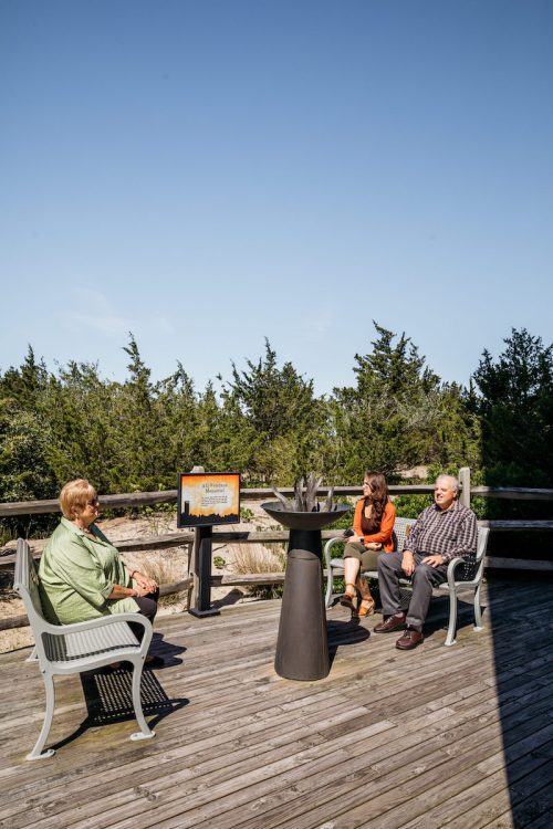 Three people sit on a wooden deck surrounded by greenery, engaged in conversation under a clear blue sky.