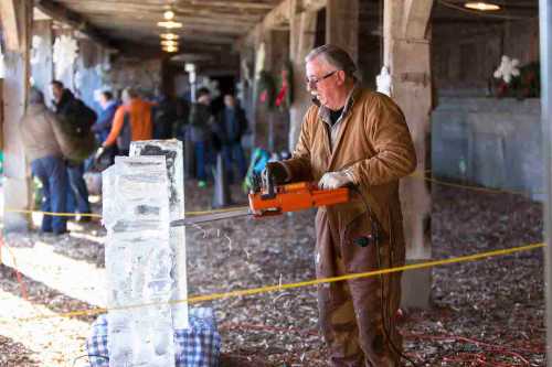 A man in a brown coat uses a chainsaw to carve an ice block in a festive indoor setting with people in the background.