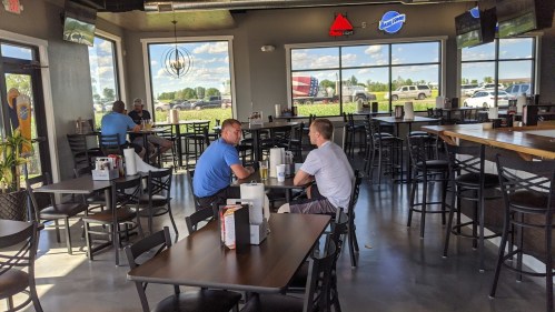 Two men sit at a table in a spacious restaurant with large windows, surrounded by empty chairs and tables.