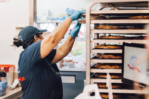 A worker in a blue shirt and gloves removes trays of baked goods from an oven in a bakery setting.