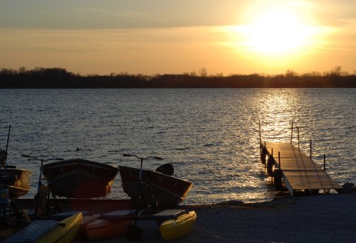 Sunset over a calm lake with boats on the shore and a wooden dock extending into the water.