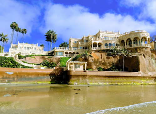 A grand coastal mansion with palm trees, perched on a cliff, overlooking a sandy beach and blue sky.